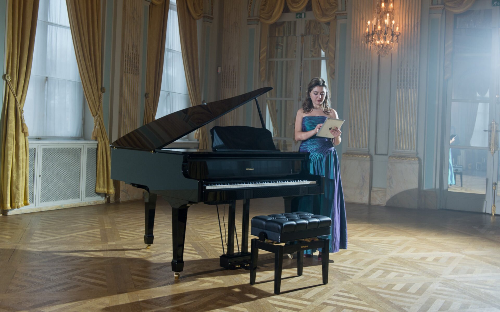 A woman in a blue dress using a tablet to connect to a Roland GP609 digital baby grand piano in a ballroom